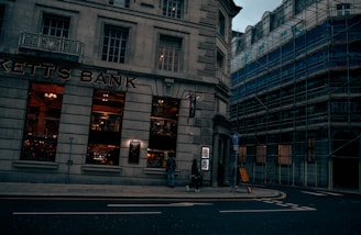 A street scene featuring a corner building with large windows, labeled as 'Kett's Bank.' The bank's windows reveal a warmly lit interior with decorative elements. Adjacent buildings are covered in scaffolding, suggesting construction or renovation work. A few pedestrians are visible on the sidewalk, and a signpost is placed near the corner.