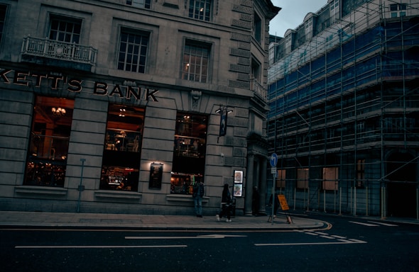 A street scene featuring a corner building with large windows, labeled as 'Kett's Bank.' The bank's windows reveal a warmly lit interior with decorative elements. Adjacent buildings are covered in scaffolding, suggesting construction or renovation work. A few pedestrians are visible on the sidewalk, and a signpost is placed near the corner.