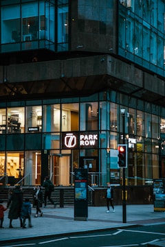 A modern building with large, illuminated glass windows. The ground floor has a sign that reads 'Park Row' and a lit interior showing various office spaces. Pedestrians are walking on the sidewalk, and a traffic light shows red.