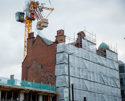A construction site featuring a brick building partially covered in scaffolding and protective sheets. An industrial crane towers above the structure, with various construction materials visible. The scene is set against a cloudy sky, creating a backdrop for the ongoing development.