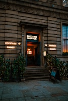 A stone building facade with a warmly lit entrance labeled 'Electric Shuffle' in neon lights. Surrounding the entrance are additional glowing signs advertising cocktails, games, brunch, and events. Steps lead up to the door, flanked by ornate iron railings adorned with greenery and string lights.