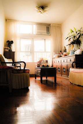 A cozy vintage-style living room with polished wooden floors and soft natural lighting. There are two armchairs with cushions on either side of a small wooden table. On the table, various small items and snacks are placed. The room's focal point is a large window with sheer curtains and an air conditioning unit above. A wooden sideboard is on the right, adorned with potted plants and decorative jars.