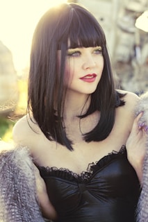 A woman with straight black hair and bangs is wearing a black strapless garment with lace detailing. She has bright makeup with green and purple eyeshadow and red lipstick, set against a background with a warm golden glow.
