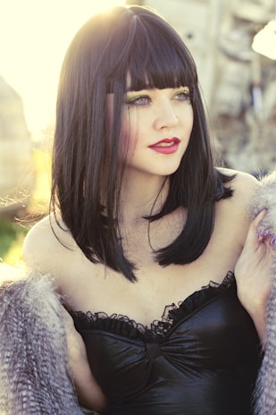 A woman with straight black hair and bangs is wearing a black strapless garment with lace detailing. She has bright makeup with green and purple eyeshadow and red lipstick, set against a background with a warm golden glow.