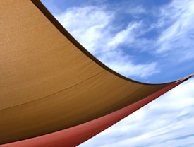 A large fabric canopy stretches across the image, casting shadows underneath. The canopy is composed of two shades, a mustard yellow and a rusty orange, with a clear blue sky and gentle white clouds visible above.
