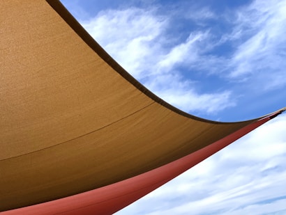A large fabric canopy stretches across the image, casting shadows underneath. The canopy is composed of two shades, a mustard yellow and a rusty orange, with a clear blue sky and gentle white clouds visible above.