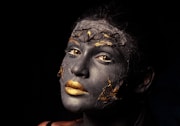 A close-up of a person's face painted in black with gold accents, resembling cracked earth. The eyes stand out with a piercing gaze, enhanced by golden eye makeup. The texture of the face paint creates an artistic and dramatic effect.