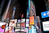 A bustling urban scene filled with brightly lit billboards and advertisements on high-rise buildings, featuring various advertisements for Broadway shows, fashion brands, and electronic products. The area is illuminated by colorful lights, with numerous signs and banners prominently displayed against the night sky.