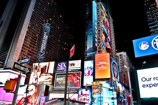 A bustling urban scene filled with brightly lit billboards and advertisements on high-rise buildings, featuring various advertisements for Broadway shows, fashion brands, and electronic products. The area is illuminated by colorful lights, with numerous signs and banners prominently displayed against the night sky.