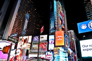 A bustling urban scene filled with brightly lit billboards and advertisements on high-rise buildings, featuring various advertisements for Broadway shows, fashion brands, and electronic products. The area is illuminated by colorful lights, with numerous signs and banners prominently displayed against the night sky.