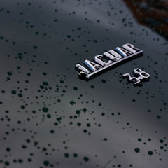A close-up view of a glossy black surface with water droplets scattered across it. The focus is on a silver emblem displaying the word 'JAGUAR' and the numbers '3.8', indicating a car model or engine specification.