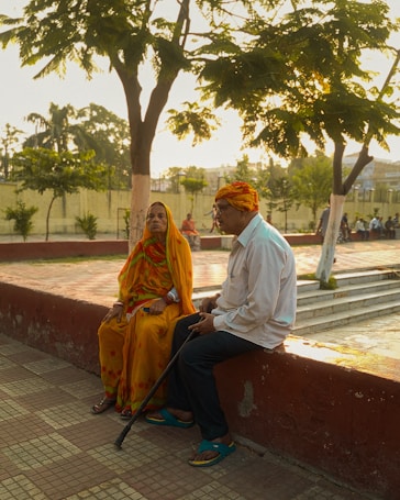 An elderly couple is seated on a low wall in a park with trees around. The woman is dressed in vibrant orange and yellow traditional clothing, while the man wears a white shirt and holds a walking stick. They appear to be engaged in a quiet conversation under the warm glow of a setting sun.