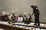 A group of people sitting on the floor against a wall, holding signs with messages like 'My faith demands ceasefire' and 'Justice' written in multiple languages. A police officer stands in front of them, interacting with the group.