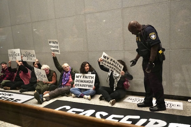 A group of people sitting on the floor against a wall, holding signs with messages like 'My faith demands ceasefire' and 'Justice' written in multiple languages. A police officer stands in front of them, interacting with the group.