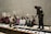 A group of people sitting on the floor against a wall, holding signs with messages like 'My faith demands ceasefire' and 'Justice' written in multiple languages. A police officer stands in front of them, interacting with the group.
