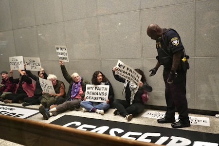 A group of people sitting on the floor against a wall, holding signs with messages like 'My faith demands ceasefire' and 'Justice' written in multiple languages. A police officer stands in front of them, interacting with the group.