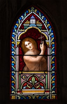 A stained glass artwork featuring a figure with golden hair holding a cross. The window is richly decorated with intricate floral and geometric patterns using vibrant colors including red, blue, and gold. The figure appears serene and is enclosed within an ornate arch surrounded by detailed motifs.