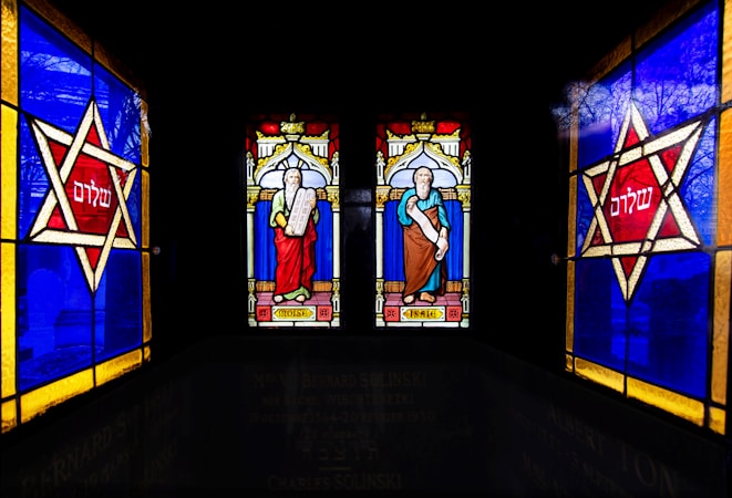 Stained glass artwork depicting two figures labeled as Moses and Isaac, each holding scrolls, surrounded by intricate designs. On either side, large red stars with Hebrew text are framed by bold blue and yellow borders.
