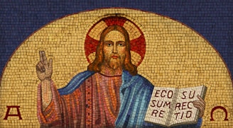A detailed mosaic depicts a figure with a halo, holding an open book with Latin text. The figure is draped in a blue and red robe, with a gesture of blessing from the right hand. The background consists of a golden aureole and a deep blue surrounding, with Greek letters Alpha and Omega on the sides.