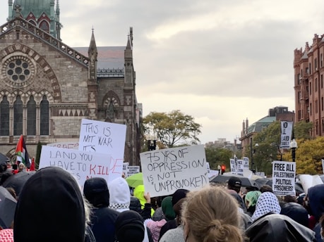 A large group of people are gathered in a protest in front of a historic building with Gothic architectural features. The crowd is holding various signs with messages advocating for political causes, some of which include phrases like 'This is not war' and 'Free all Palestinian political prisoners'. The scene takes place outside with overcast skies, and some trees are visible in the background.