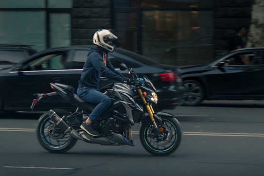 A person wearing a helmet and a hoodie is riding a black motorcycle on the street. The motorcycle appears to be in motion, and there are several cars in the background, including a black sedan. The scene is set in an urban environment, possibly near a building with large windows.