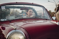 A person sits behind the wheel of a classic red car, with a focused expression. The car has stickers on the windshield, including one that reads 'Gulf.' The background shows an urban setting with trees and buildings.