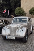 A vintage silver car is parked on a cobblestone street. The car is well-preserved, showcasing chrome details and classic round headlights. Behind the car, two Georgian flags are visible, along with trees and a stone building.