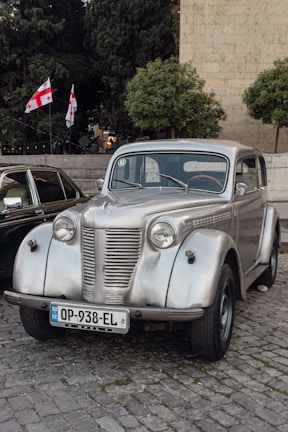 A vintage silver car is parked on a cobblestone street. The car is well-preserved, showcasing chrome details and classic round headlights. Behind the car, two Georgian flags are visible, along with trees and a stone building.