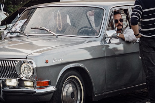 A vintage gray car with a polished exterior is parked in an outdoor setting. The car's right front window is open, and a man wearing sunglasses and a striped shirt is sitting inside, looking out. Another person is standing nearby, partially visible, wearing a striped sweater. A paper or pamphlet is visible on the car's windshield.