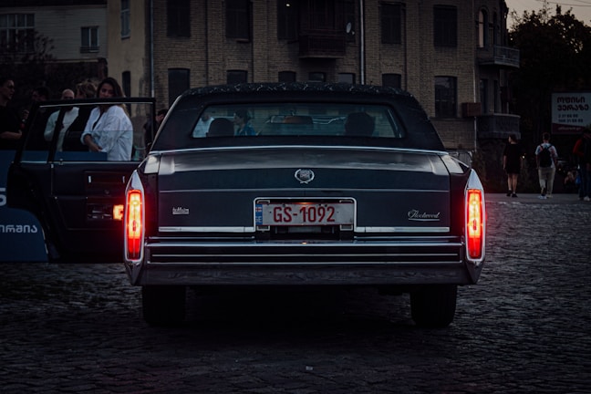 A vintage black Cadillac with distinctive taillights is parked on a cobblestone street. The car's license plate reads 'GS-1092'. Several people are visible in the background, with one person standing by an open car door as others walk on the sidewalk. The setting is urban with a few buildings visible, creating a moody atmosphere.