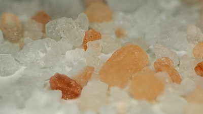 A close-up view of a mixture of coarse salt and pink Himalayan salt crystals. The salt grains vary in size and texture, with some appearing rough and jagged. The image has a soft focus, concentrating on the subtle details of the salt crystals.