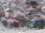 A close-up view of soap bubbles with iridescent surfaces displaying a spectrum of colors. The bubbles are connected in a cluster, creating an intricate pattern of shapes. Light reflects off the surfaces, highlighting subtle textures and variations in color.