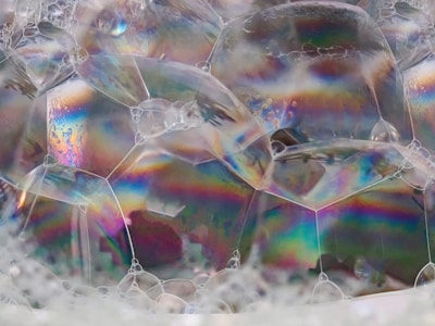 A close-up view of soap bubbles with iridescent surfaces displaying a spectrum of colors. The bubbles are connected in a cluster, creating an intricate pattern of shapes. Light reflects off the surfaces, highlighting subtle textures and variations in color.