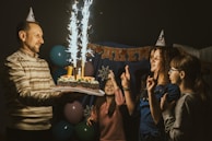 A celebration scene with a man holding a cake adorned with sparklers, surrounded by two women and a girl wearing party hats. Balloons are visible in the background along with a festive banner.