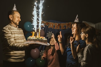 A celebration scene with a man holding a cake adorned with sparklers, surrounded by two women and a girl wearing party hats. Balloons are visible in the background along with a festive banner.