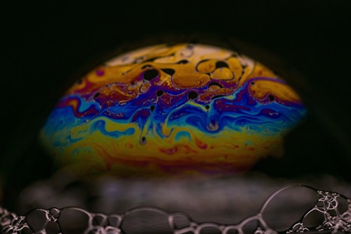 A close-up of a soap bubble showing a vibrant array of swirling colors including blue, yellow, red, and purple. The surface of the bubble displays intricate patterns resembling abstract art, with dark spots and fluid line formations. Surrounding the bubble, delicate soap film encases smaller bubbles, creating a fascinating contrast between the colorful center and the monochromatic edges.