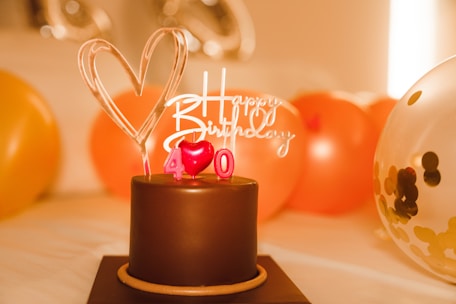 A chocolate cake adorned with a heart-shaped topper and red candles forming the number 40. A 'Happy Birthday' sign is also present on top of the cake. Surrounding the cake are balloons in various colors, including orange and a transparent one with confetti inside. The background is warmly lit, giving a celebratory atmosphere.