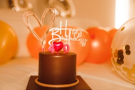 A chocolate cake adorned with a heart-shaped topper and red candles forming the number 40. A 'Happy Birthday' sign is also present on top of the cake. Surrounding the cake are balloons in various colors, including orange and a transparent one with confetti inside. The background is warmly lit, giving a celebratory atmosphere.