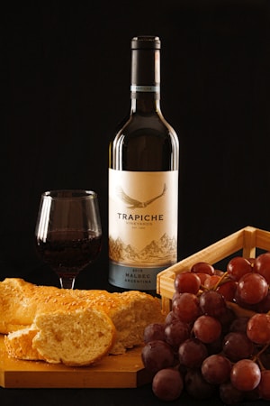 A bottle of Trapiche vineyard Malbec wine is centered alongside a glass of red wine. Nearby, a loaf of sliced bread and a cluster of red grapes add to the setting, all resting on a wooden surface against a dark background.