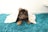 A small puppy lies on a plush teal rug, partially covered by a white blanket adorned with colorful animal prints. The puppy has dark fur with lighter brown markings, and its ears are perked up.