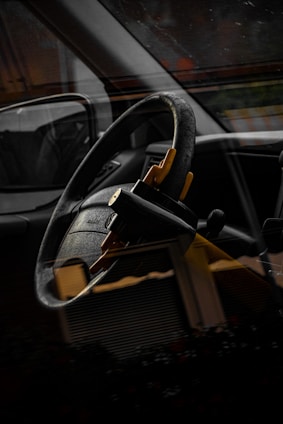 A close-up of a car interior showing a steering wheel with a yellow steering wheel lock attached, reflected through the car window. The image is somewhat dark, highlighting the black and yellow colors.