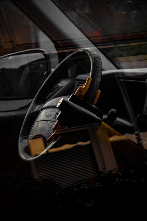 A close-up of a car interior showing a steering wheel with a yellow steering wheel lock attached, reflected through the car window. The image is somewhat dark, highlighting the black and yellow colors.