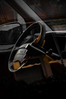 A close-up of a car interior showing a steering wheel with a yellow steering wheel lock attached, reflected through the car window. The image is somewhat dark, highlighting the black and yellow colors.