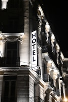 A well-lit hotel building at night, showcasing ornate architectural details and a prominent illuminated sign that reads 'HOTEL' with three stars below it. The facade features decorative moldings, balconies with wrought iron railings, and spotlights illuminating the classical details.