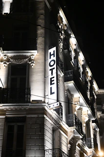 A well-lit hotel building at night, showcasing ornate architectural details and a prominent illuminated sign that reads 'HOTEL' with three stars below it. The facade features decorative moldings, balconies with wrought iron railings, and spotlights illuminating the classical details.