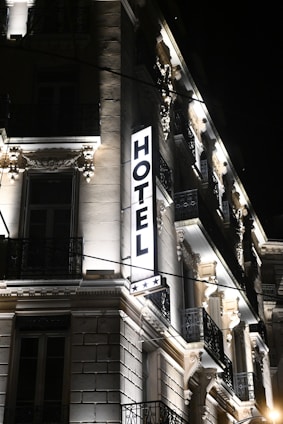 A well-lit hotel building at night, showcasing ornate architectural details and a prominent illuminated sign that reads 'HOTEL' with three stars below it. The facade features decorative moldings, balconies with wrought iron railings, and spotlights illuminating the classical details.