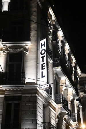 A well-lit hotel building at night, showcasing ornate architectural details and a prominent illuminated sign that reads 'HOTEL' with three stars below it. The facade features decorative moldings, balconies with wrought iron railings, and spotlights illuminating the classical details.