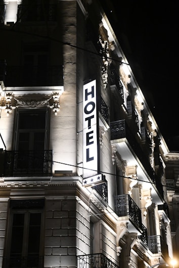 A well-lit hotel building at night, showcasing ornate architectural details and a prominent illuminated sign that reads 'HOTEL' with three stars below it. The facade features decorative moldings, balconies with wrought iron railings, and spotlights illuminating the classical details.