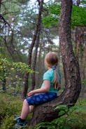 A child with long hair sits on a curved tree trunk in a lush, green forest. The child is wearing a turquoise shirt, blue shorts with patterns, and sneakers. The forest is dense with tall, slender trees and vibrant green foliage, creating a serene and natural setting.