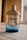 A glass bottle with a beige cap and a transparent blue lower section, featuring the brand name 'KAIAK' printed in blue. The bottle is positioned on a light wooden surface with blurred background elements visible through a window.